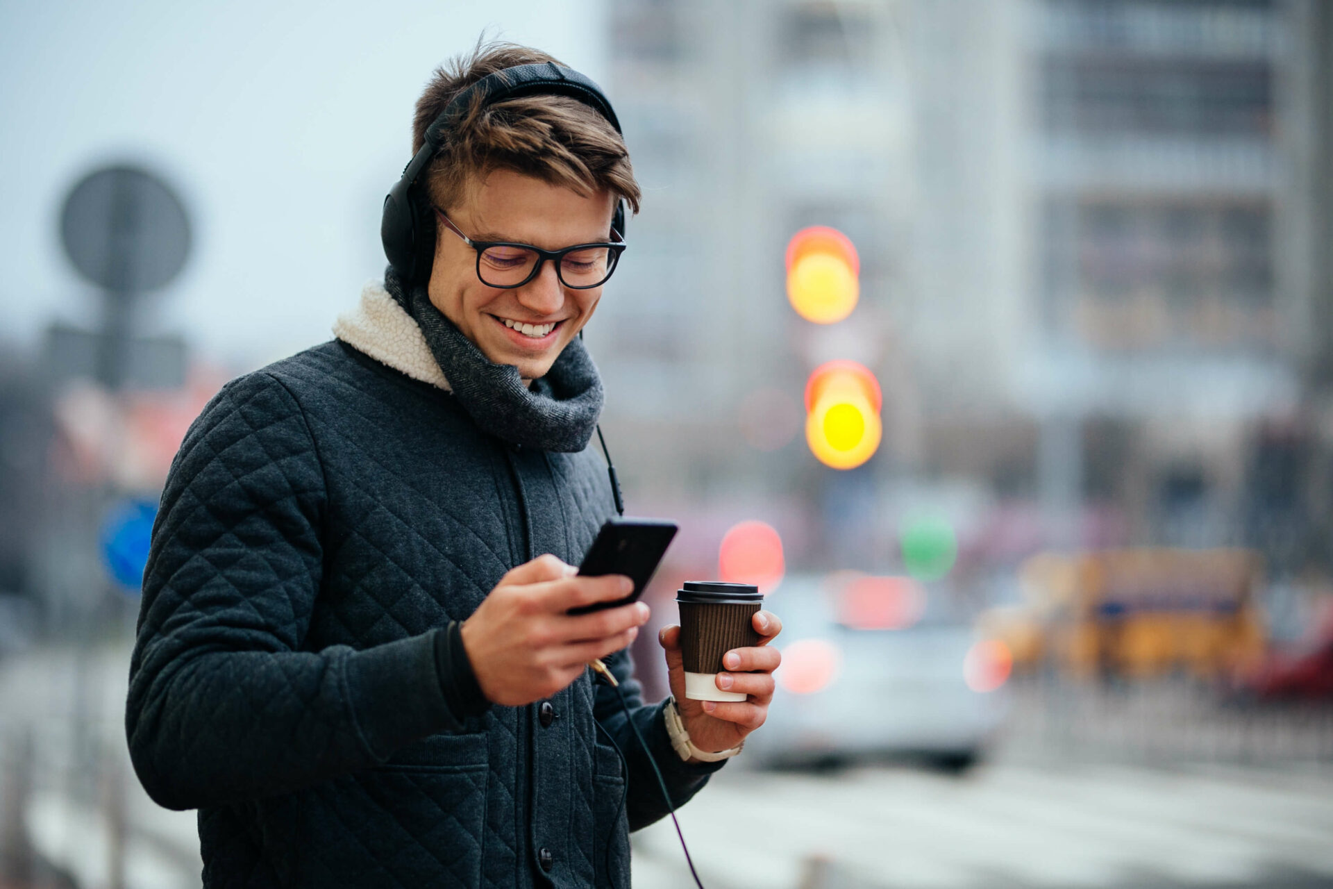 Smiling young man wearing headphones and a winter jacket, holding a coffee cup while looking at his phone on a city street.