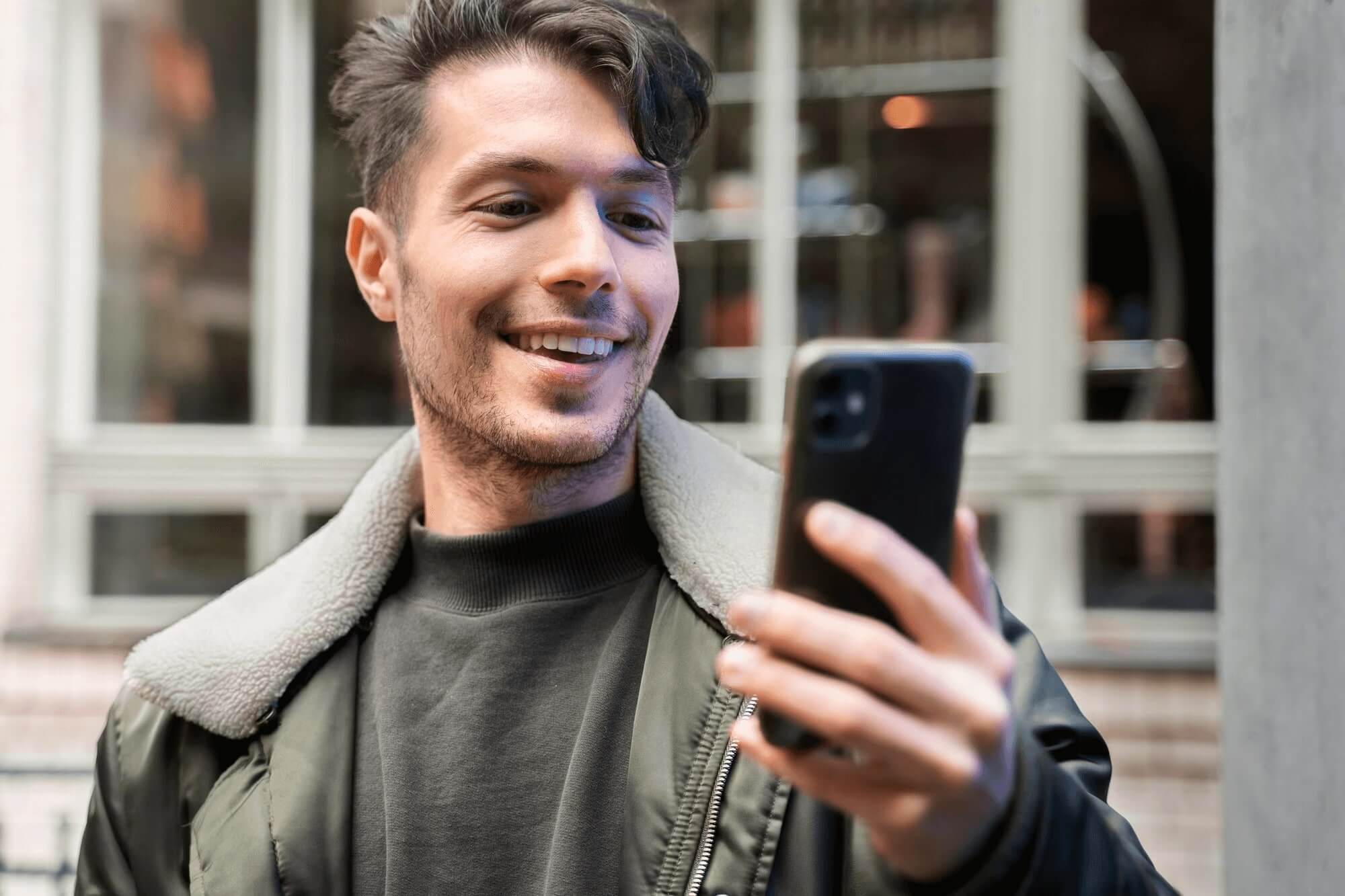 Smiling young man in a casual jacket looking at his phone outdoors, appearing engaged in a conversation or message.