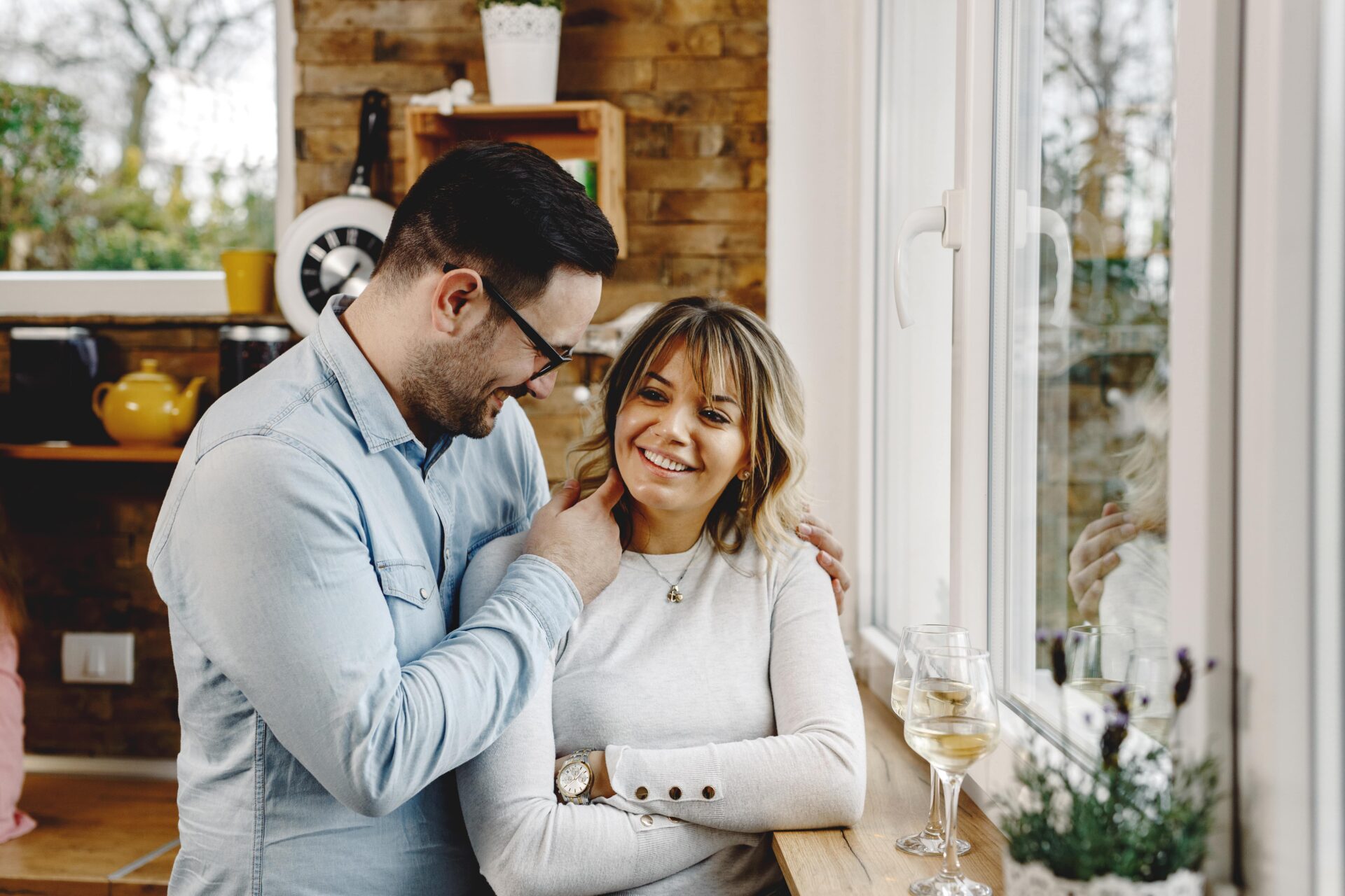 Happy couple smiling and bonding together in a cozy kitchen by the window with wine glasses